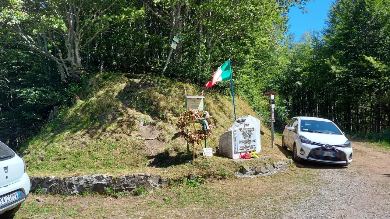 Monumento commemorativo con bandiera italiana e corona d'alloro al passo delle radici, circondato da alberi e con un'auto parcheggiata.