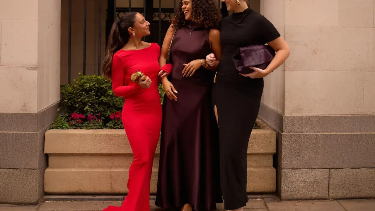 Three women in elegant gowns, discussing what not to wear to a wedding. One in red, one in plum, and one in black.