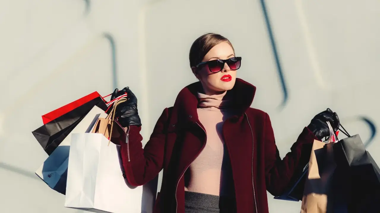 Donna con occhiali da sole e rossetto rosso, avvolta in un cappotto bordeaux, porta borse della spesa, simbolo di moda veloce.