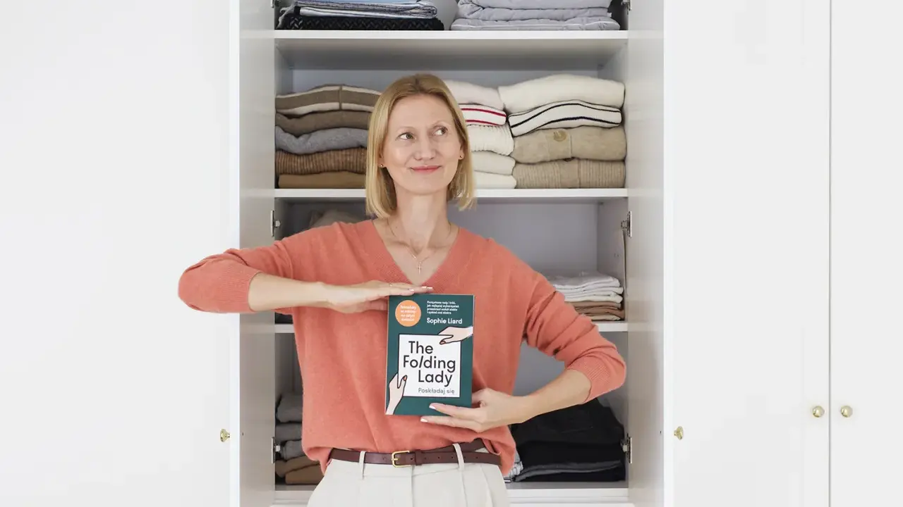 Kobieta z książką "The Folding Lady" pokazuje, jak składać koszulki, stojąc przed otwartą szafą pełną schludnie ułożonych ubrań.