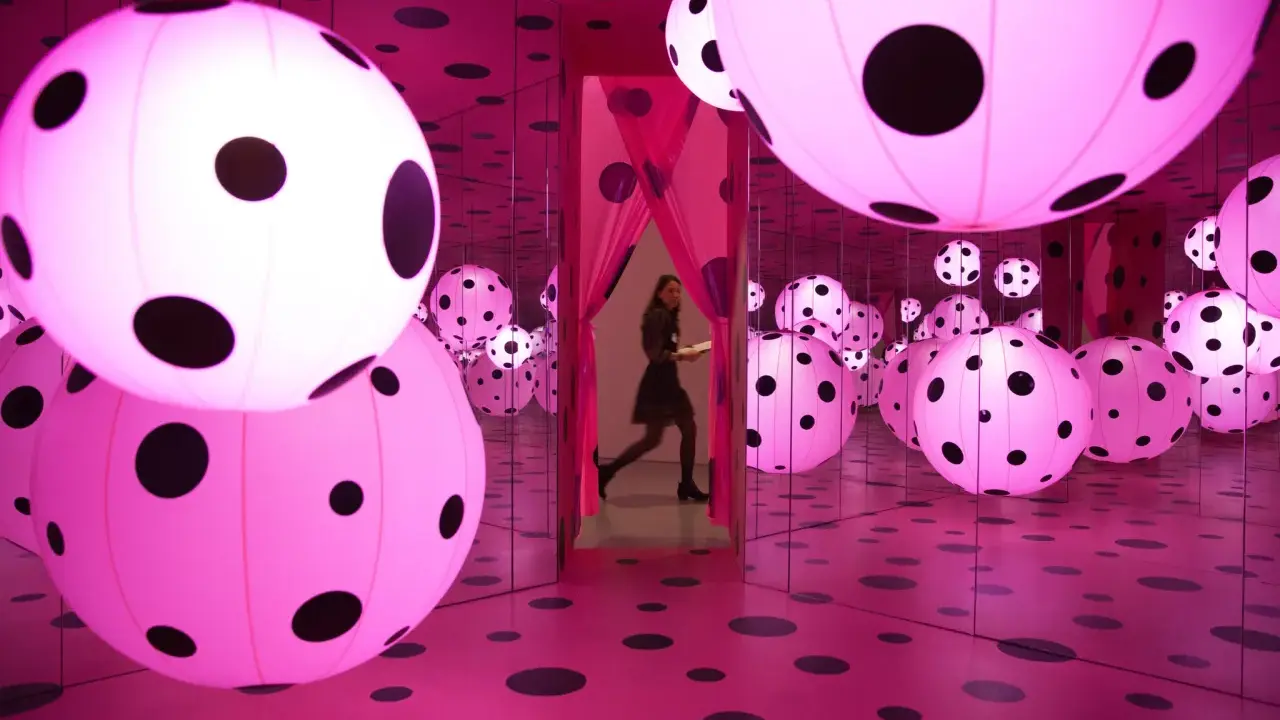 A woman walks through a mirrored room filled with pink polka-dotted spheres, a signature of Yayoi Kusama's immersive art.