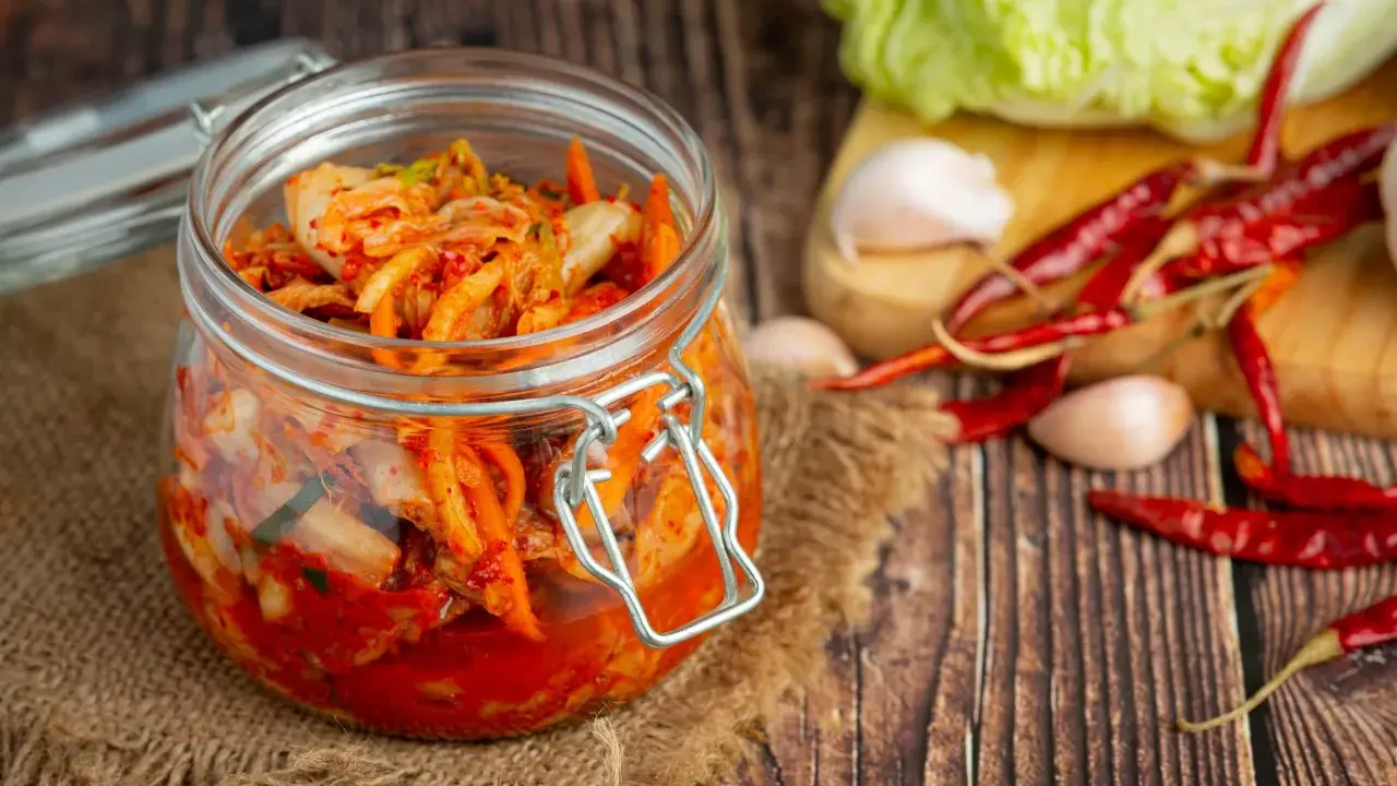 Słoik z kimchi na tle desek, obok kapusta, czosnek i papryczki chili.