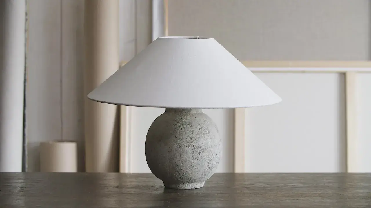 Bordslampa med rund, texturerad fot av lufttorkande lera, målad i grågröna toner. En vit lampskärm sitter ovanpå.