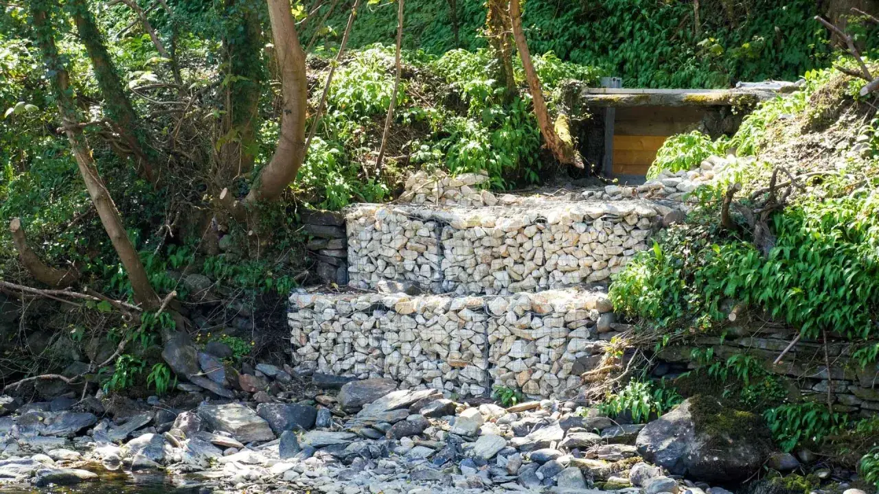 Estructura de muro de gavion con piedras blancas, rodeada de vegetación y rocas junto a un río.
