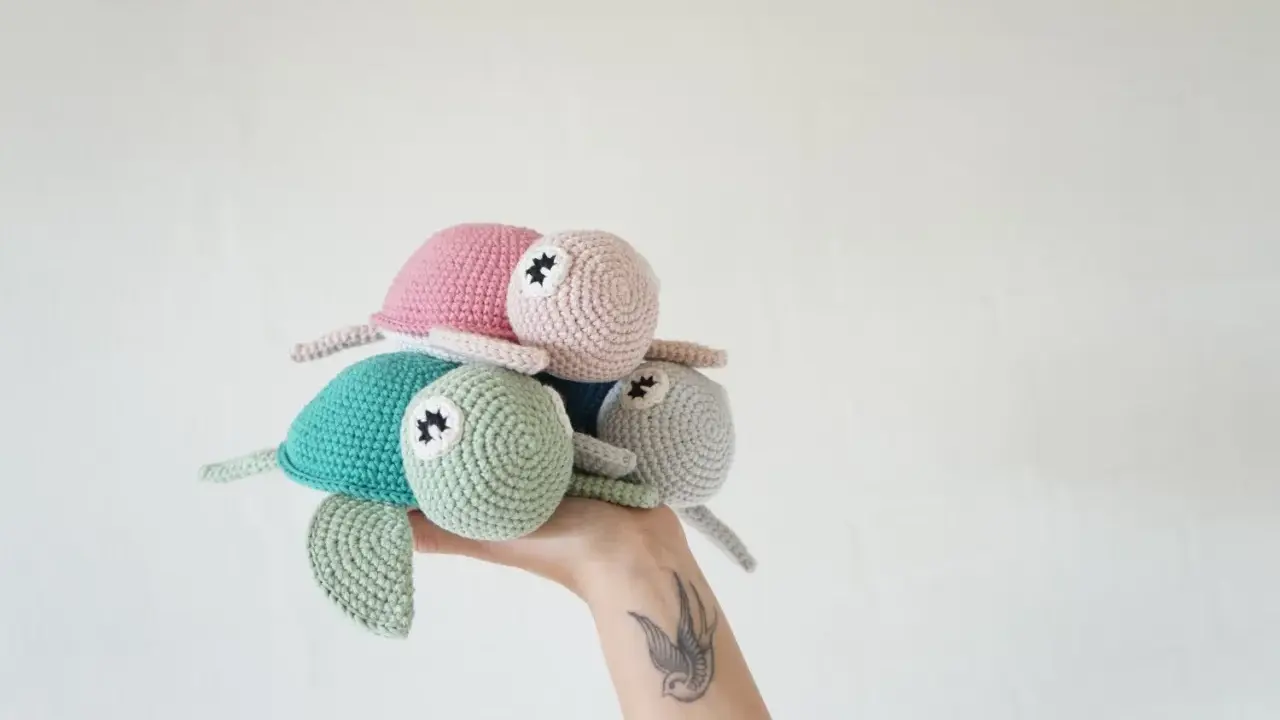 Una mano sostiene un trío de adorables tortugas a crochet: una verde menta, una rosa y una gris claro.
