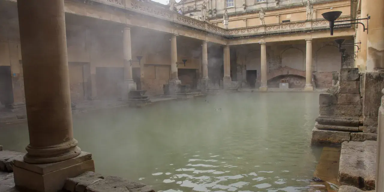 Dampf steigt aus dem Becken der römischen Therme auf. Säulen und Bögen umgeben das Wasser, in dem Enten schwimmen.