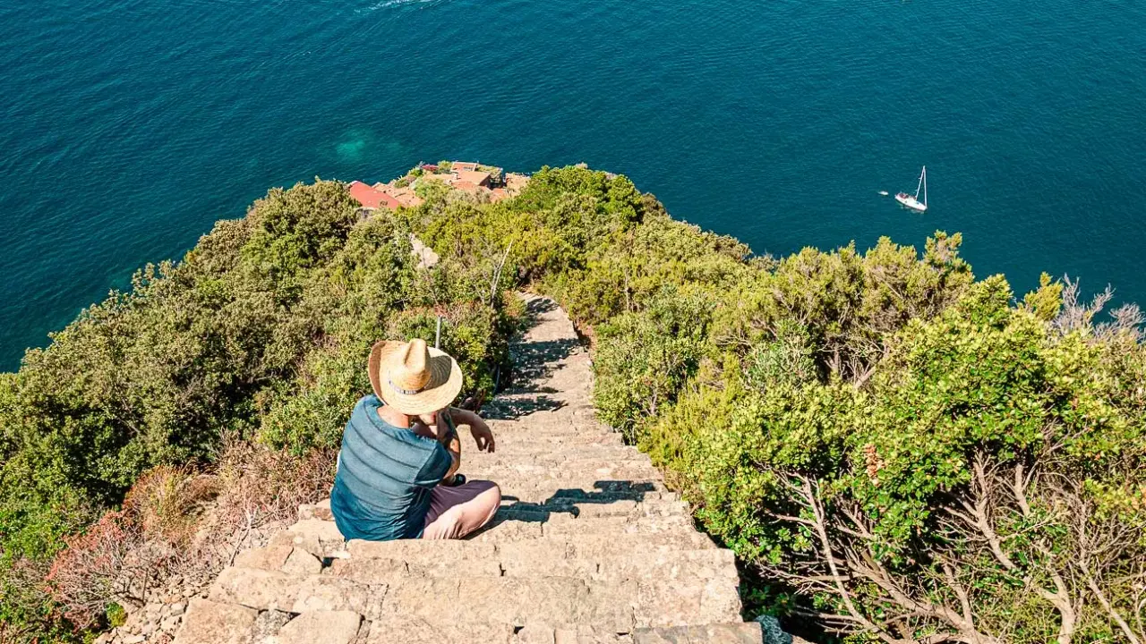 Un uomo con cappello di paglia seduto su una scalinata di pietra guarda il mare blu. Un sentiero scende verso il mare, suggerendo come arrivare a Monesteroli.