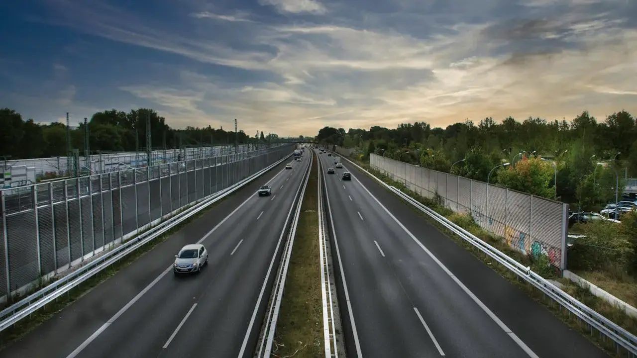 Szeroka autostrada z samochodami w oddali, otoczona zielenią i barierami dźwiękochłonnymi. Jazda autostradą pod pochmurnym niebem.