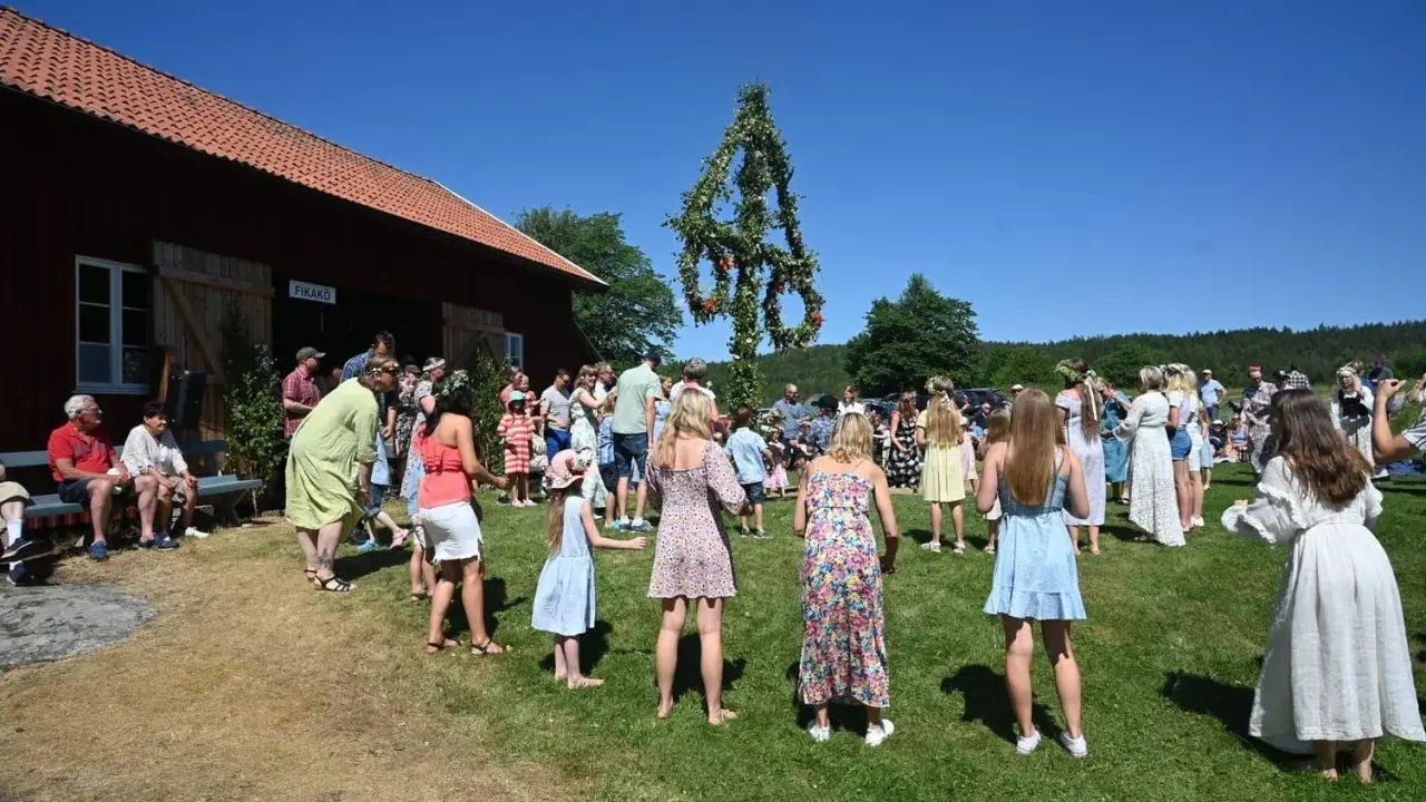 Folk dansar runt midsommarstången på midsommar afton. En vacker sommardag med klarblå himmel.