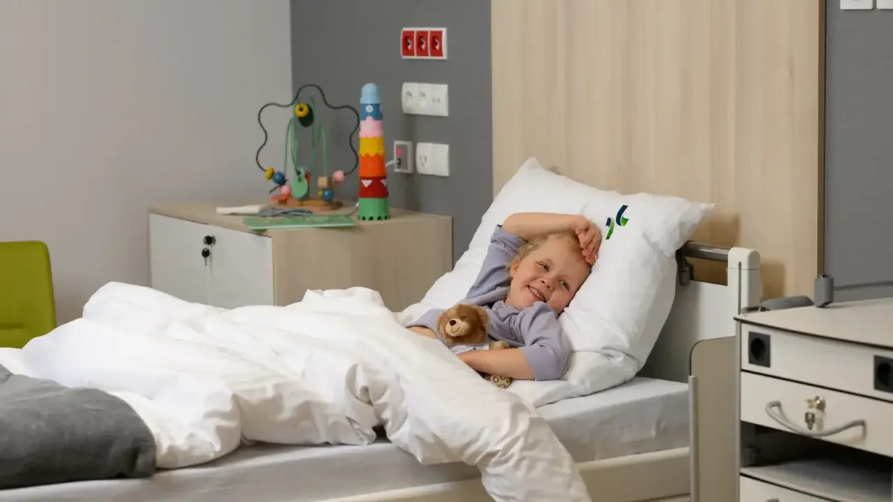 Uśmiechnięte dziecko w szpitalnym łóżku, przytulające misia. Obok zabawka edukacyjna i szafka.