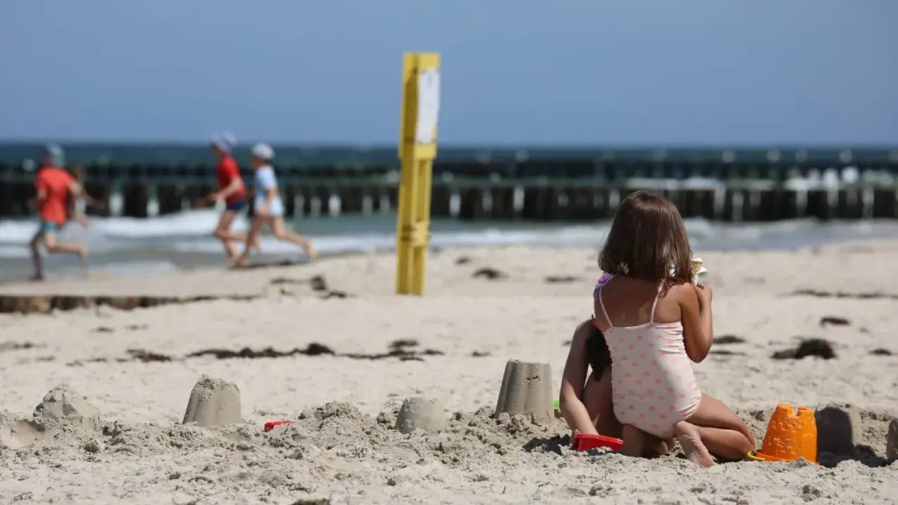Dzieci budują zamki z piasku na plaży, w tle widać morze i falochrony. Dźwirzyno molo w oddali.