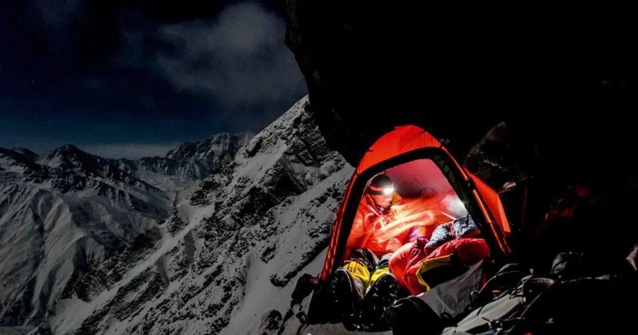 Nocny obóz na zboczu himalajskiego parbatu. W pomarańczowym namiocie rozświetlonym czołówką odpoczywa alpinista, otoczony surowym, ośnieżonym krajobrazem.