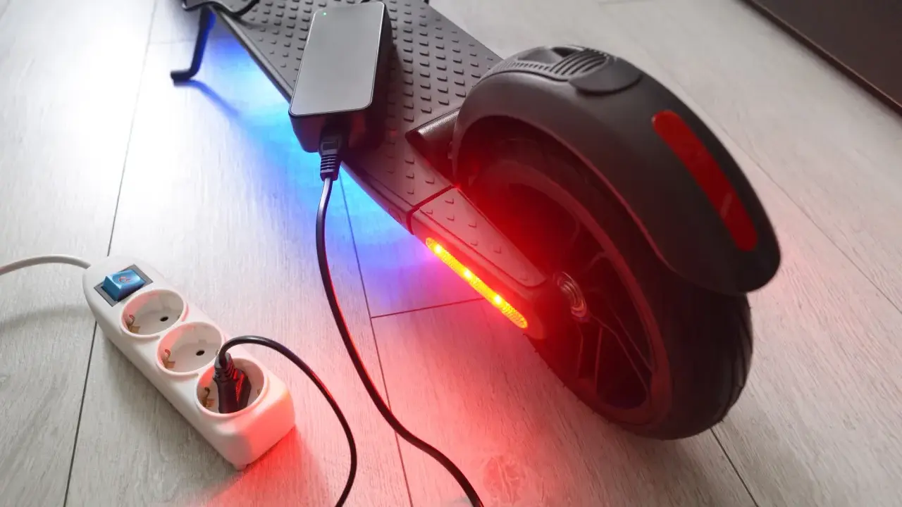 Un chargeur pour trottinette électrique est branché sur une multiprise. La trottinette est éclairée par des LED bleues et rouges.