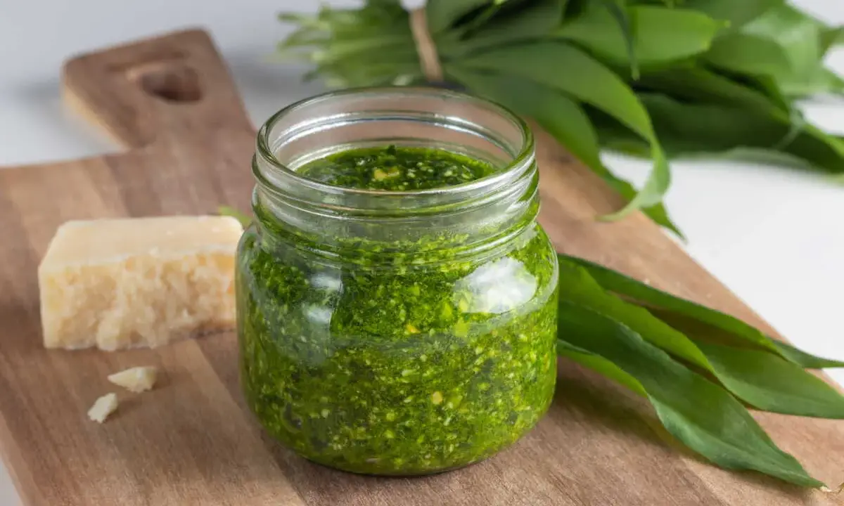 Pesto ail des ours fait maison dans un bocal, avec du fromage et des feuilles fraîches sur une planche en bois.