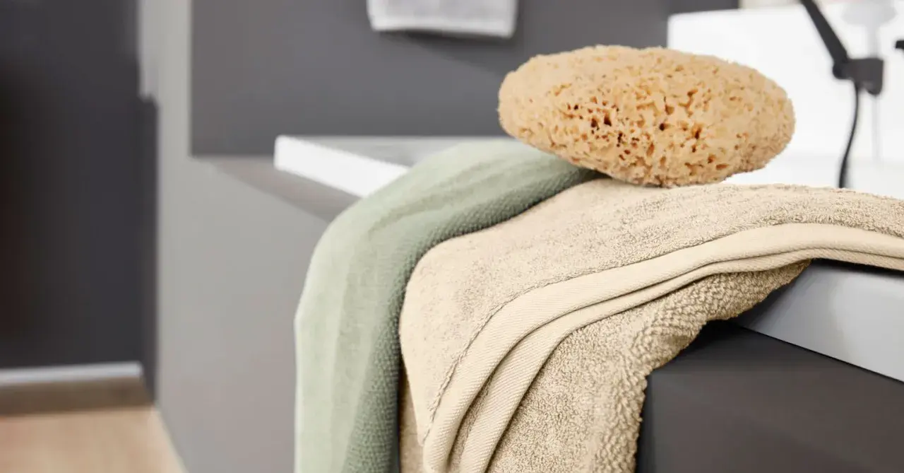 Weiche bad textilien in sanften Farben, darunter ein hellgrünes und ein beigefarbenes Handtuch, liegen neben einem natürlichen Schwamm auf der Kante einer Badewanne.