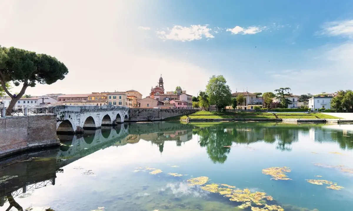 Ponte di Tiberio e il fiume Marecchia a Rimini a settembre, con riflessi sull'acqua e un cielo azzurro con nuvole bianche.