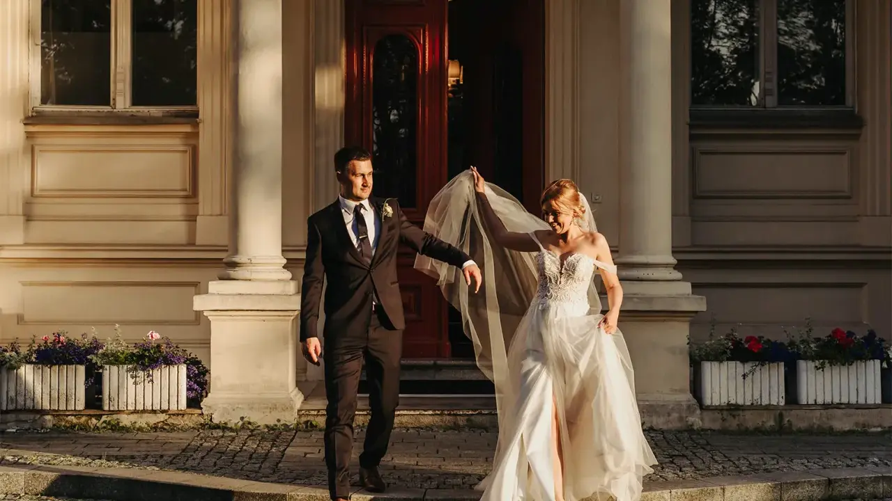 Para młoda podczas sesji zdjęciowej ślubnej schodzi po schodach przed eleganckim budynkiem. Panna młoda w zwiewnej sukni unosi welon, pan młody w garniturze patrzy na nią z uśmiechem.