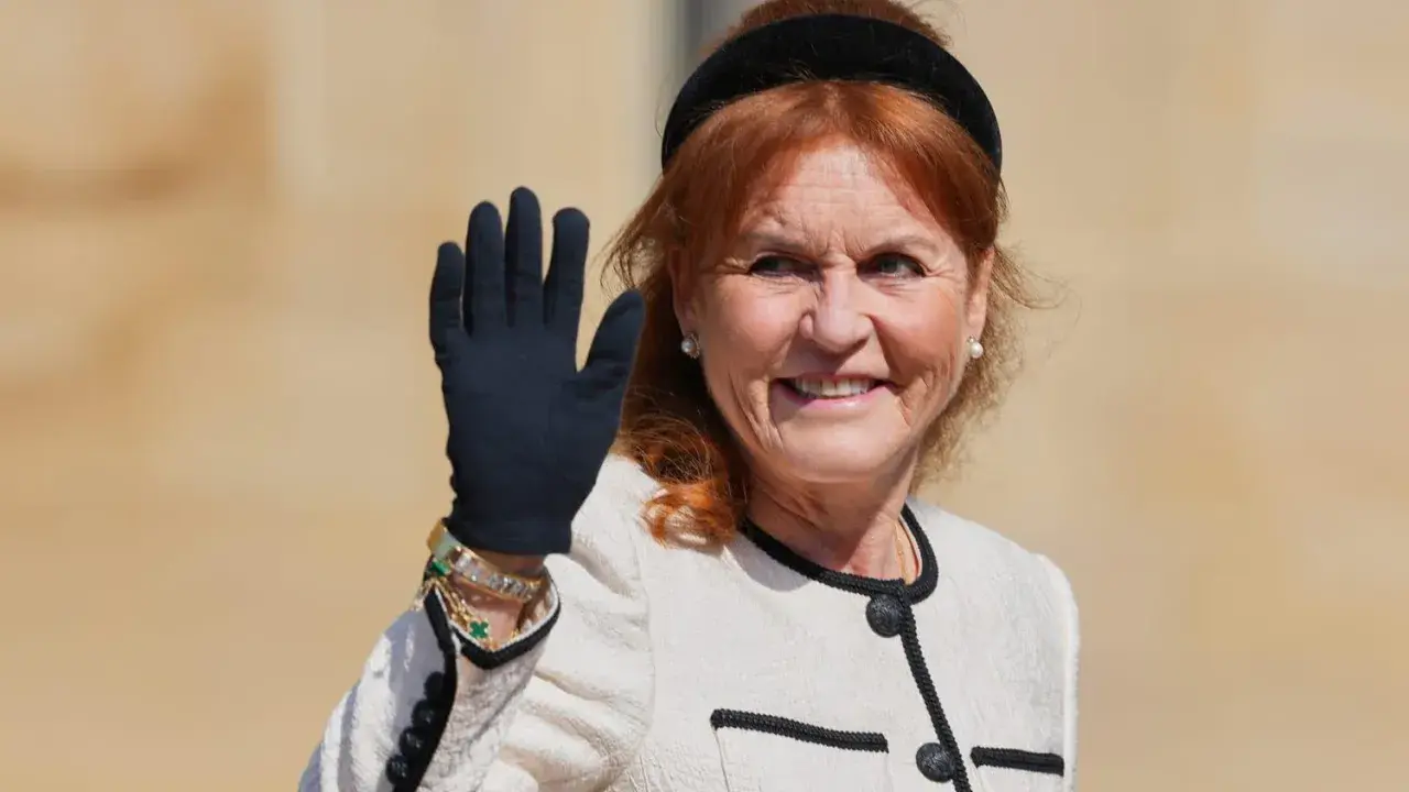 Sarah, hertiginna av York, vinkar glatt med en svart handske på sig. Hon bär en elegant ljus jacka med svarta detaljer och smycken.
