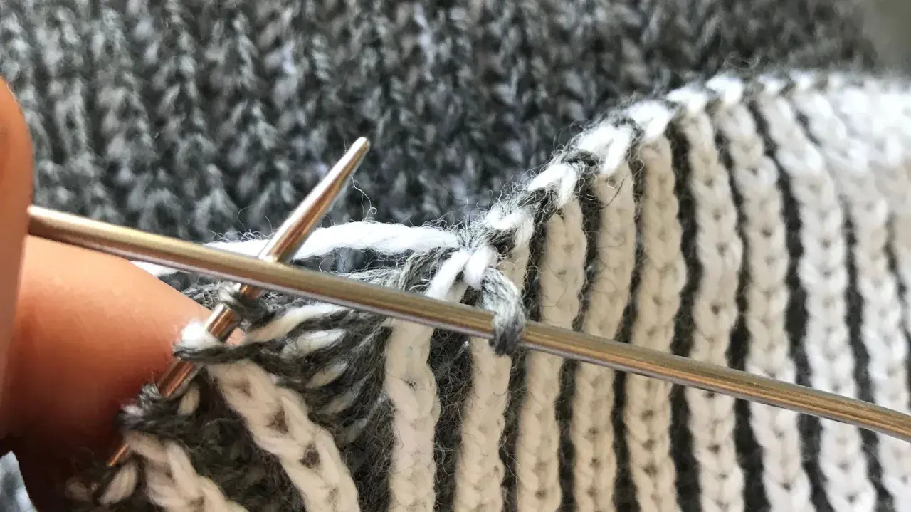 Närbild på patentstickning på rundsticka med grått och vitt garn. En tumme håller i stickan.