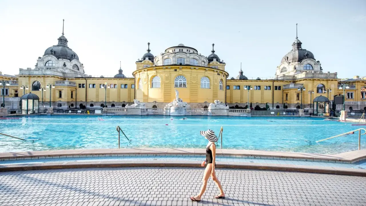 Frau mit Hut spaziert am Beckenrand des Budapester Thermalbads. Das beeindruckende gelbe Gebäude mit Kuppeln bildet die Kulisse.