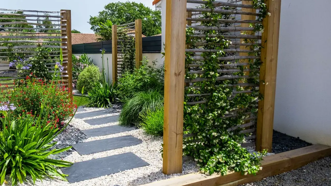 Un chemin de dalles grises serpente à travers un jardin luxuriant. Des treillages en bois servent de support à une plante brise vue grimpante, créant un espace intime.