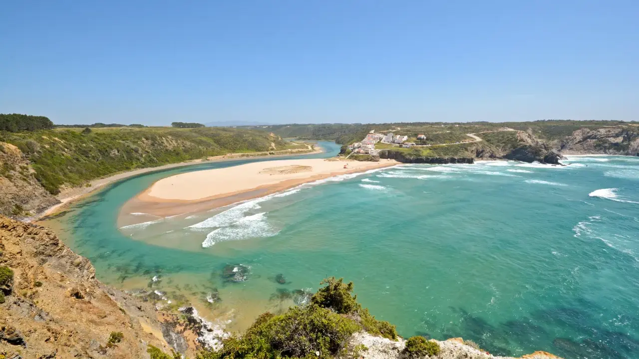 Atemberaubende Aussicht auf Praia de Odeceixe, wo ein Fluss auf den Ozean trifft. Grüne Hügel und ein weißes Dorf säumen die Küste.