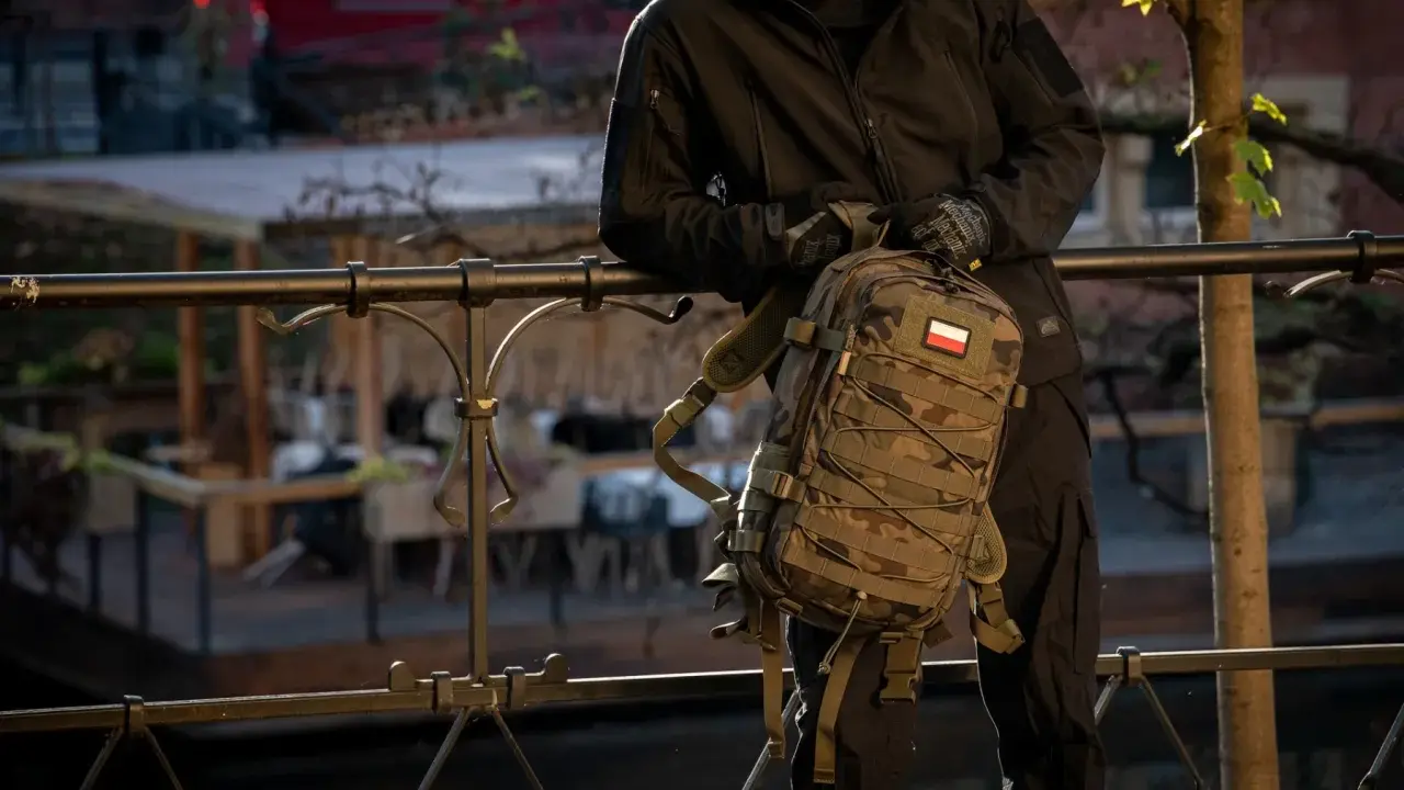 Mężczyzna w czarnej kurtce i rękawiczkach trzyma plecak w kamuflażu, z polską flagą. To jeden z wielu stylowych modeli marki plecaków.