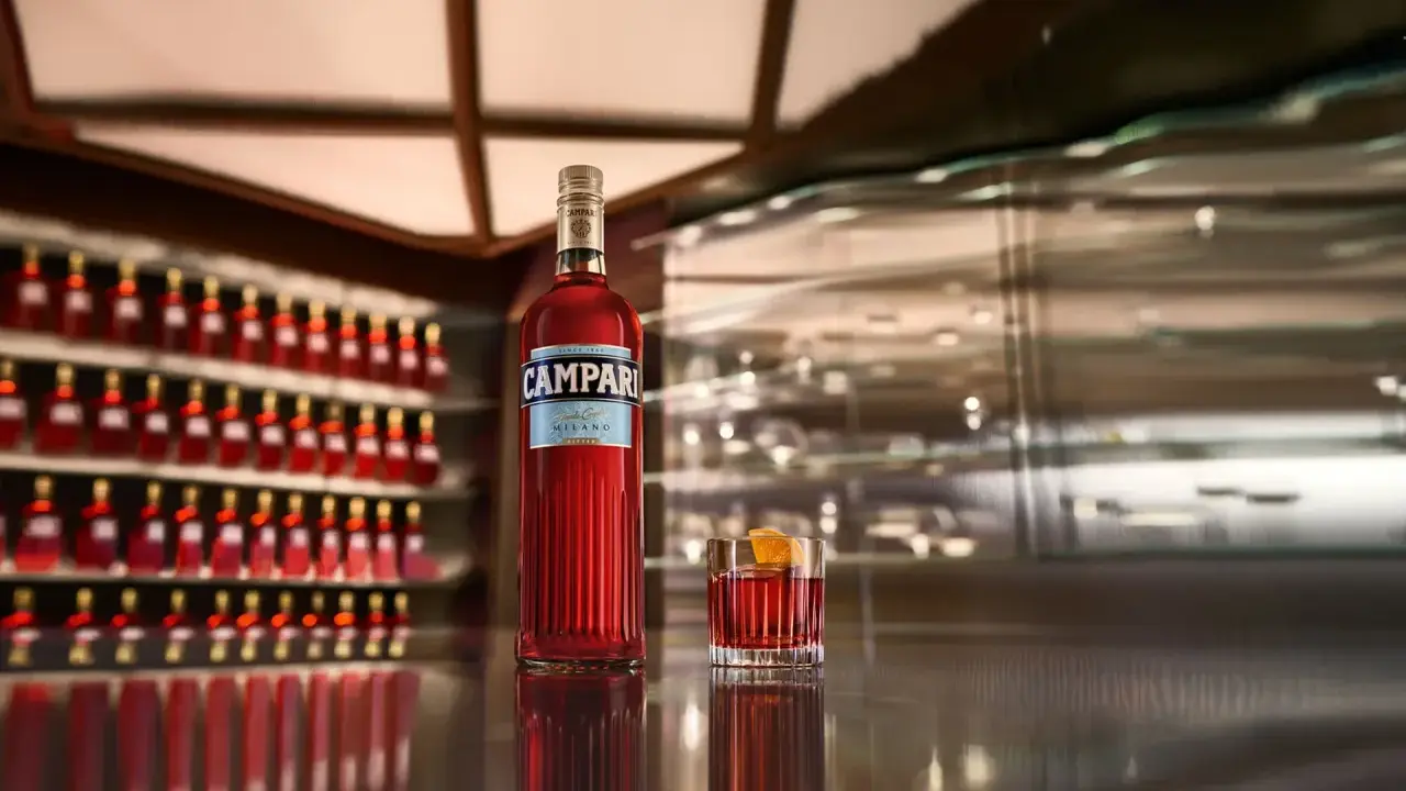 Eine Flasche Campari und ein Glas mit dem roten Aperitif stehen auf einer glänzenden Theke. Die Frage "wie schmeckt Campari?" wird durch den Anblick des ikonischen Getränks aufgeworfen.