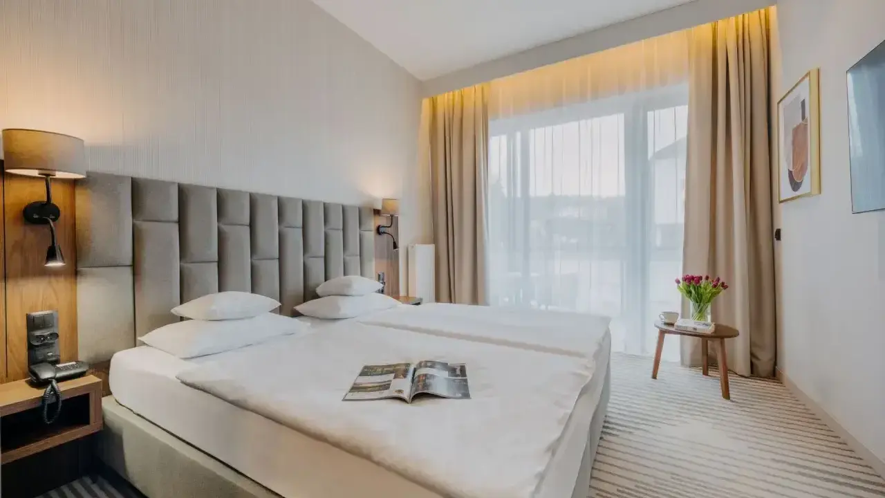 Przytulny executive room z dwoma wygodnymi łóżkami, lampkami nocnymi i dużym oknem z zasłonami. Na stoliku bukiet tulipanów.