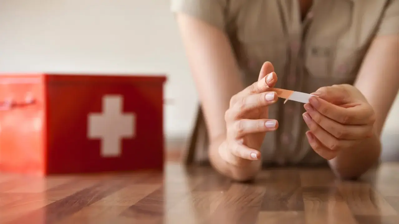 Une personne applique un pansement sur son doigt, près d'une trousse de secours rouge. L'objectif est une cicatrisation rapide.