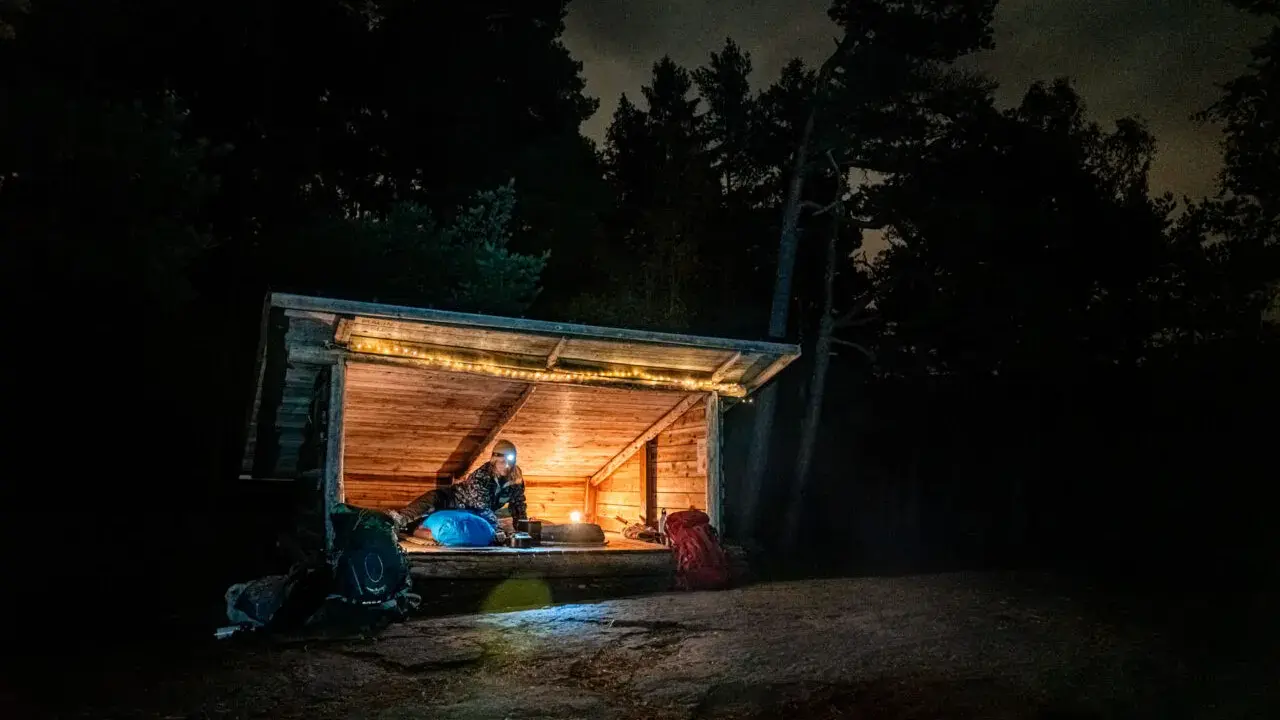 Person myser i ett upplyst vindskydd i skogen på natten. Ryggsäckar står utanför.