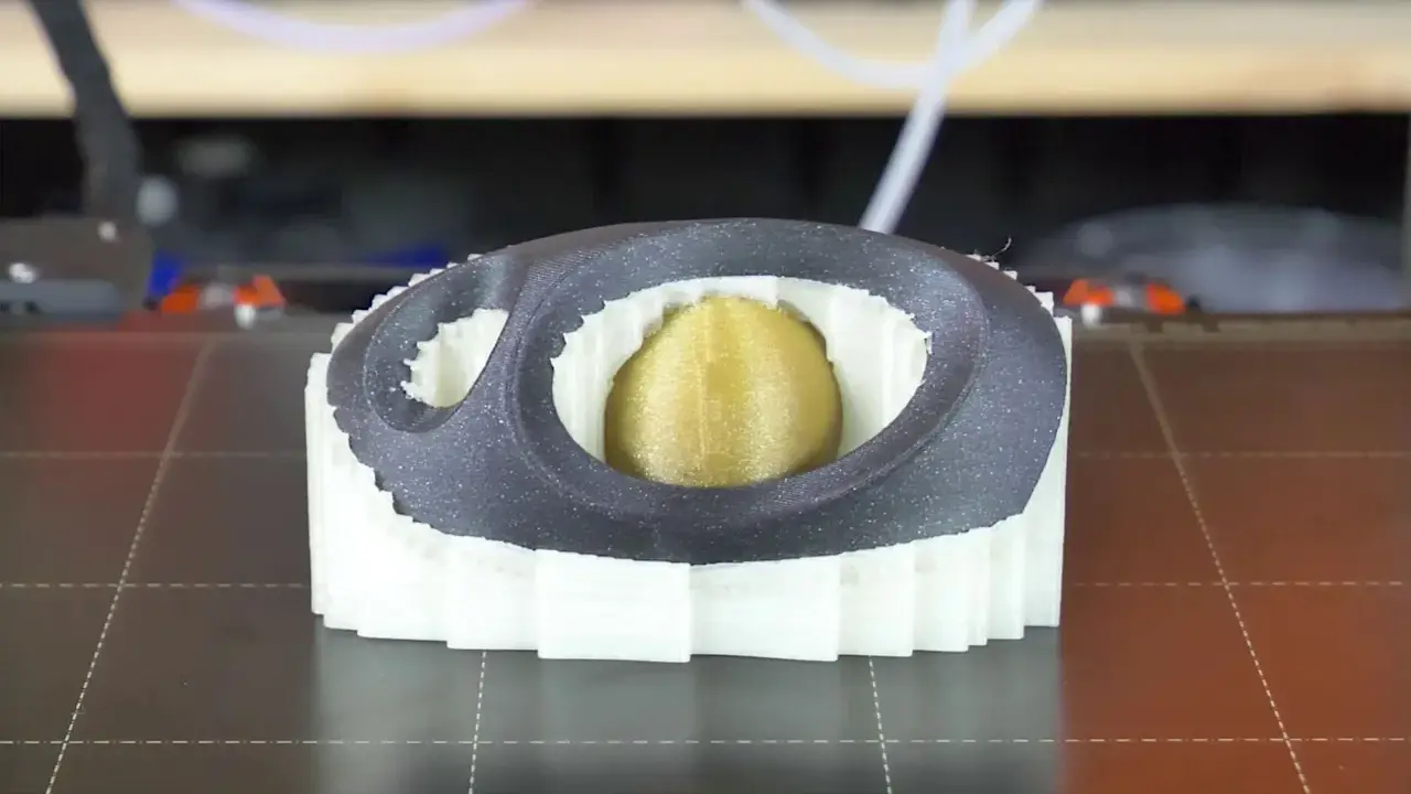 Stampa 3D di un oggetto con nucleo dorato, circondato da strati bianchi e una cornice esterna grigia. Il materiale di supporto PVA è visibile nei dettagli.