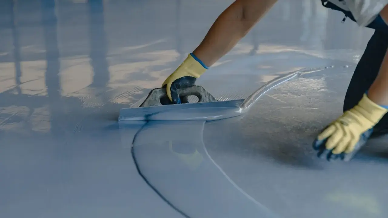 Persona con guantes amarillos aplica una capa de pintura azul clara sobre un suelo de terrazo con una llana.