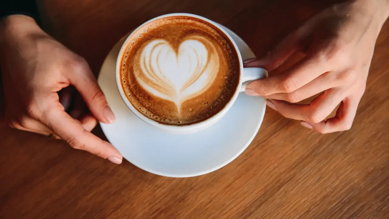 Eine Tasse Milchkaffee mit Herz-Latte-Art, gehalten von zwei Händen. Ein perfekter Start in den Tag, was ist Milchkaffee?