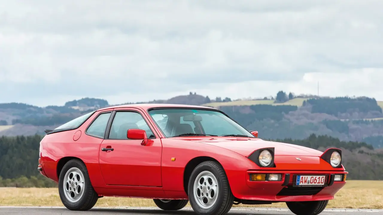 Roter Porsche 925 mit ausklappbaren Scheinwerfern steht auf einer Straße, im Hintergrund bewaldete Hügel.
