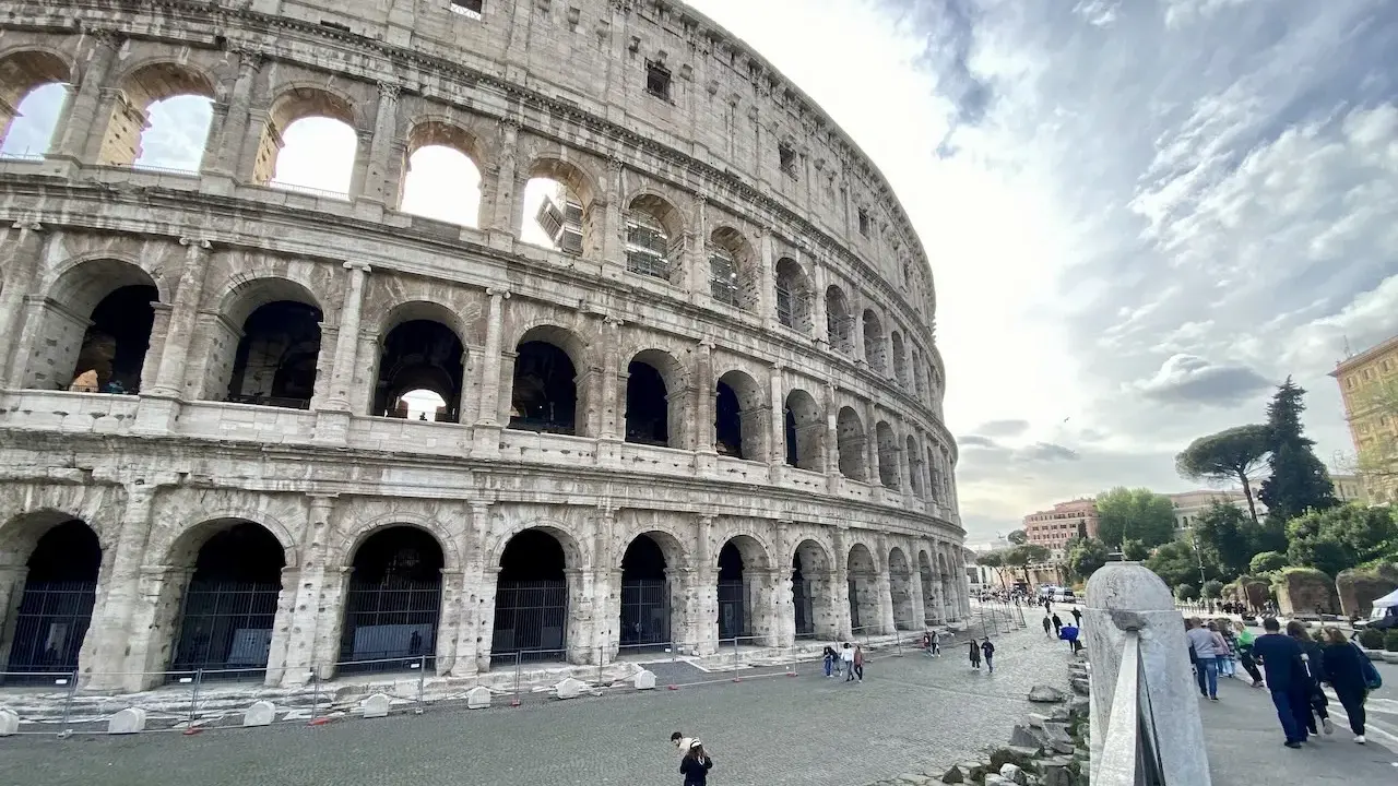 Le Colisée, monument emblématique de Rome, se dresse majestueusement sous un ciel nuageux. Des visiteurs explorent ses environs pavés.