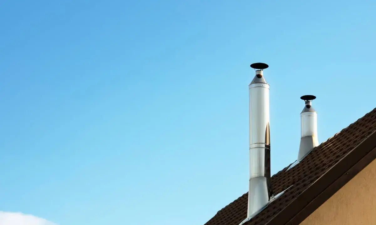 Dos tubos de chimenea metálicos se alzan sobre un tejado marrón contra un cielo azul claro. El sol calienta el metal, listo para aprovechar el calor.