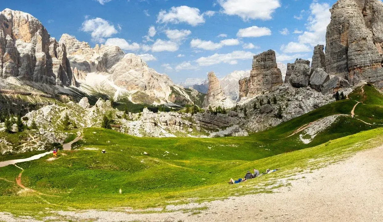 Un paesaggio mozzafiato nelle Dolomiti con le iconiche 5 torri che si ergono maestose. Un prato verde brillante invita al relax, con persone che riposano sotto il cielo azzurro.