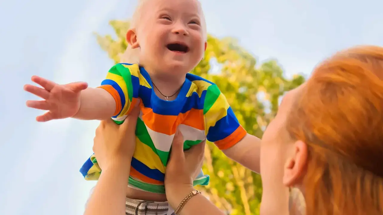 Niño con síndrome de Down, sonriente, es levantado por una persona. Sus rasgos son visibles y llenos de alegría.