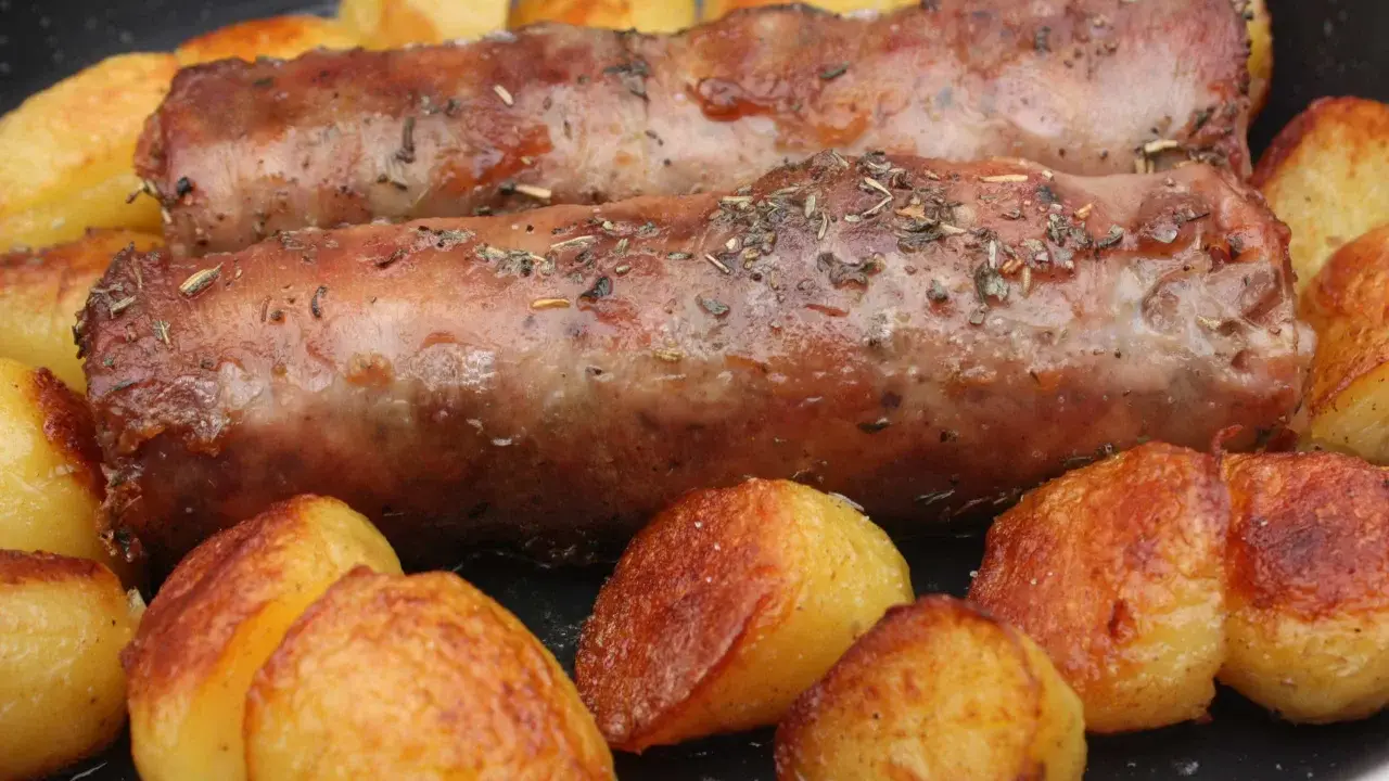 Deux andouillettes artisanales dorées, parsemées d'herbes, mijotent avec des pommes de terre rôties dans une poêle.