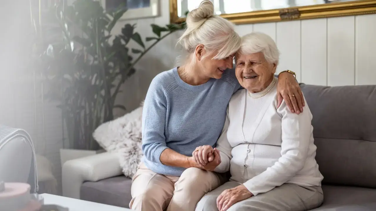 Deux femmes âgées s'enlacent tendrement sur un canapé. L'une d'elles semble visiter sa mère, une belle dame, dans sa maison en EHPAD.
