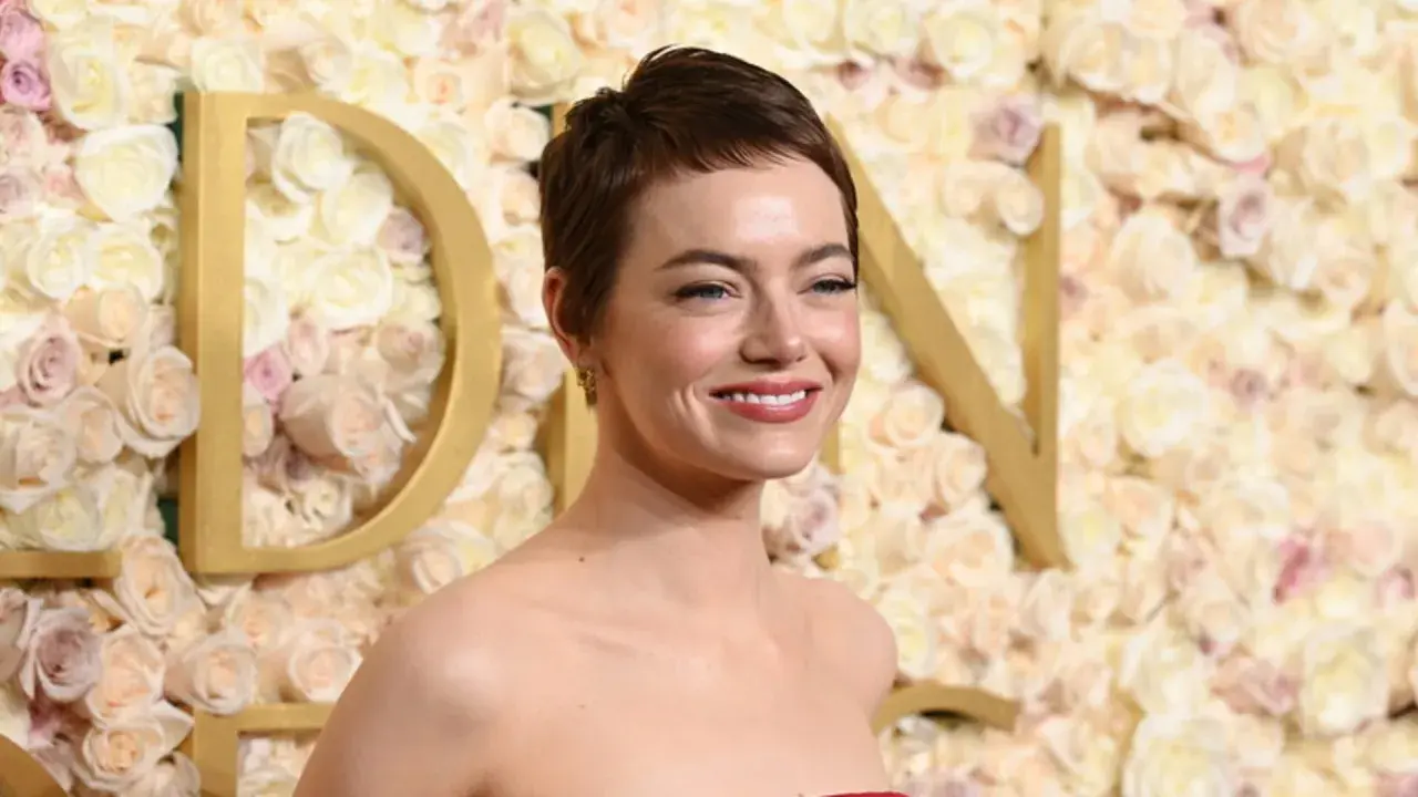 Emma Stone sorride, con un trucco che esalta i colori che stanno bene alle castane con pelle chiara. Lo sfondo è un muro di rose rosa e bianche.