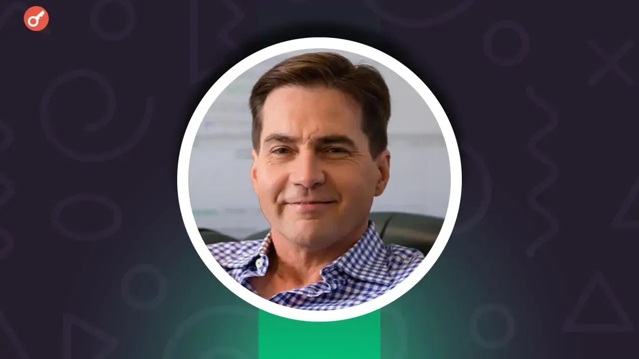Uśmiechnięty Craig Wright w niebiesko-białej koszuli w kratę, z kluczem w lewym górnym rogu.