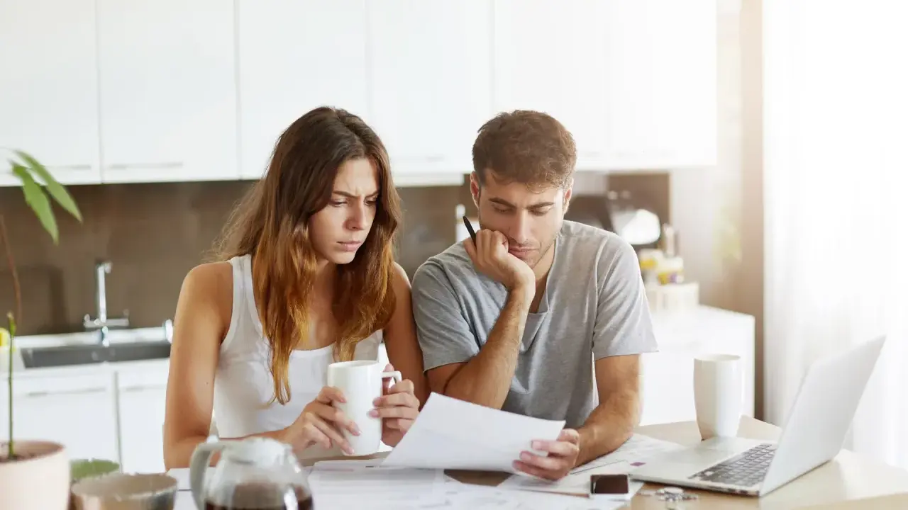 Couple pensif devant des papiers, peut-être une attestation d'assurance villégiature, dans une cuisine lumineuse.