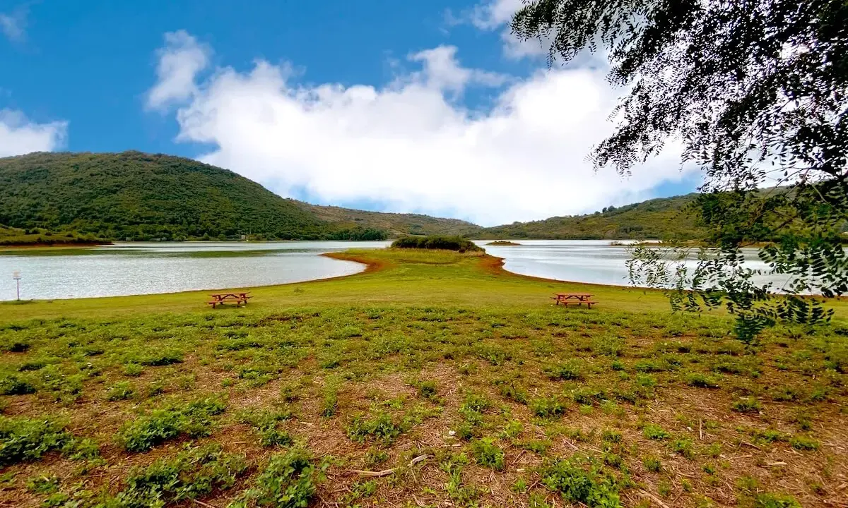 Prato verde con tavoli da picnic affacciati sul **lago di Canterno**, circondato da dolci colline sotto un cielo azzurro con nuvole bianche.
