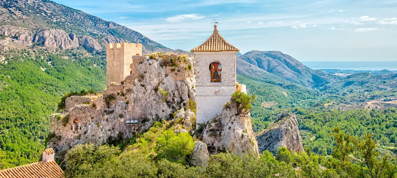 Das Castell de Guadalest thront auf einem Felsen, umgeben von grünen Bergen und dem blauen Meer in der Ferne. Ein Glockenturm ragt empor.