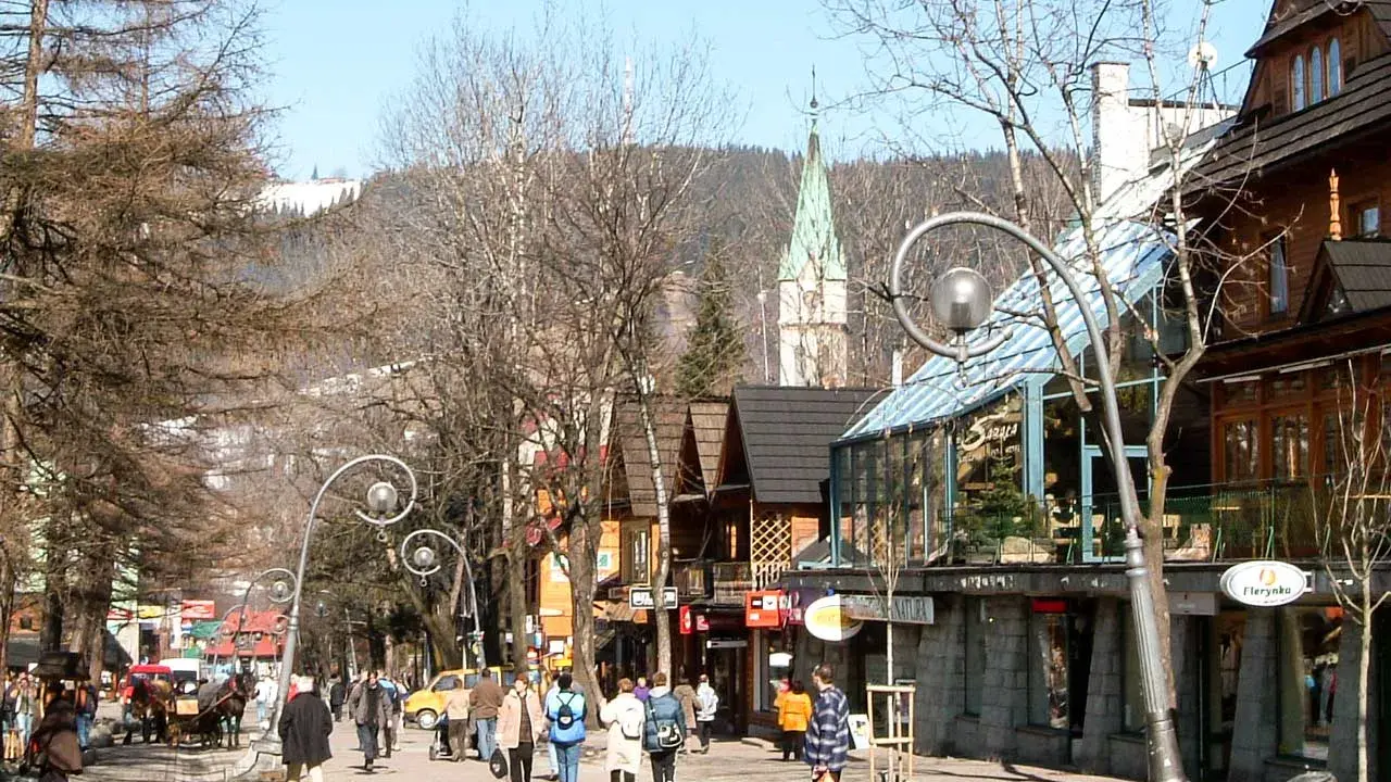 Spacer po urokliwej ulicy Krupówki w Zakopanem. Ludzie przechadzają się, podziwiając góralską architekturę i widok na Tatry.