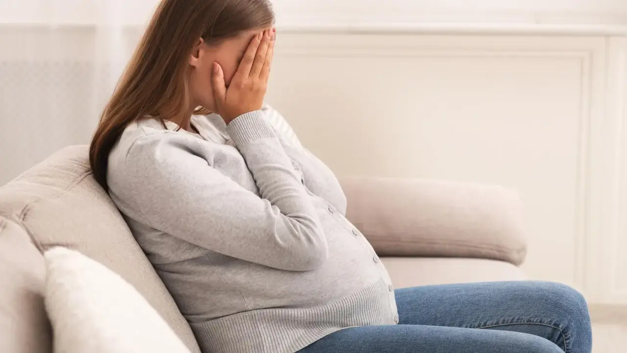 Kobieta w ciąży siedzi na kanapie, zakrywając twarz dłońmi. Jej postawa wyraża głęboki smutek, być może związany z diagnozą wady letalnej.