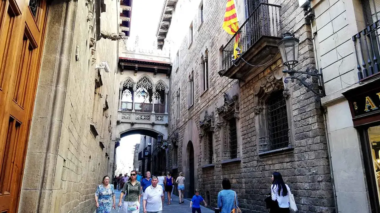 Menschen schlendern durch eine enge Gasse in der **Barcelona Altstadt**. Über ihnen spannt sich eine historische Brücke mit filigranen Bögen.