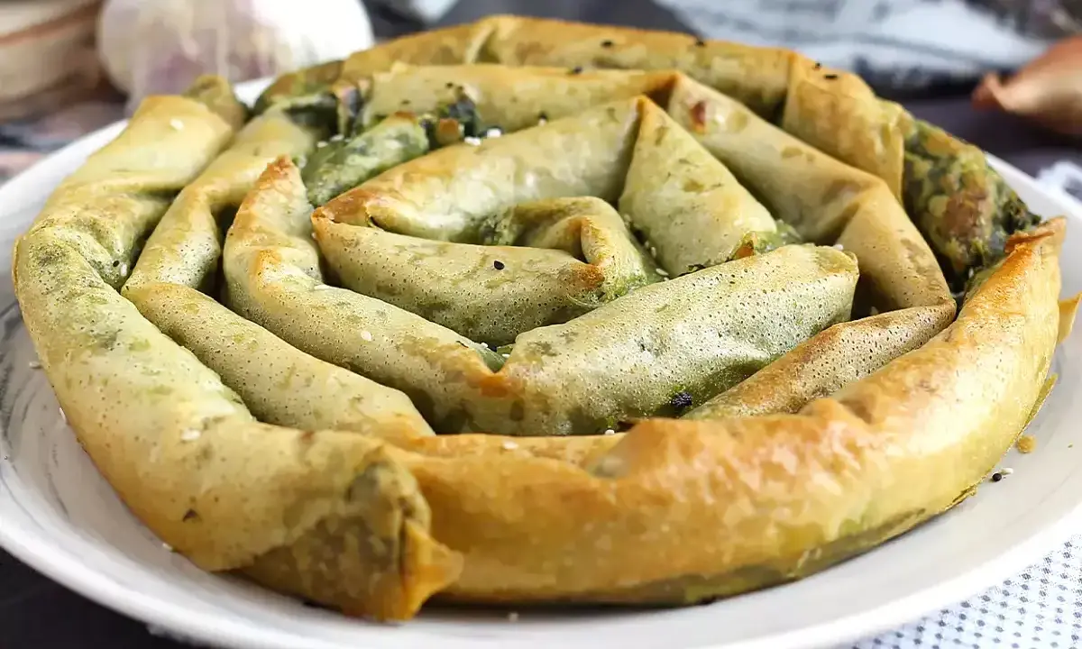 En gyllene, spiralformad burek med spenatfyllning, perfekt för ett hemlagat burek recept. Vitlök och lök i bakgrunden.
